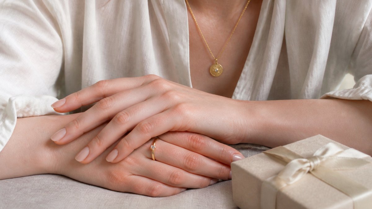 Close-up of a woman's hands wearing a delicate gold pendant necklace and a simple ring, resting on a linen surface beside a small gift box