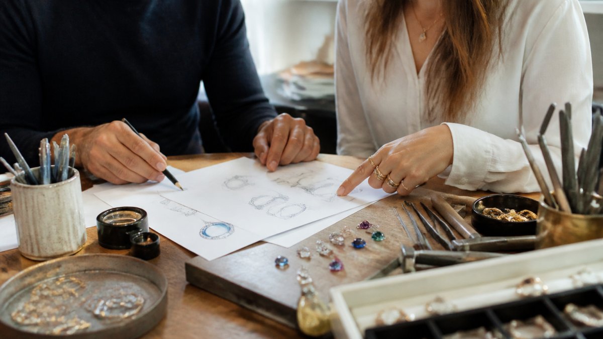 A jeweler and a client reviewing sketched jewelry designs and loose gemstones at a bright wooden workbench, with hand tools and a loupe nearby