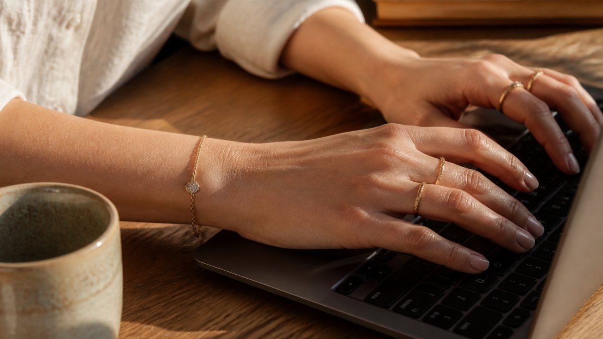 Woman wearing delicate gold rings and a bracelet from ethical jewelry brands while typing at a sunlit desk.